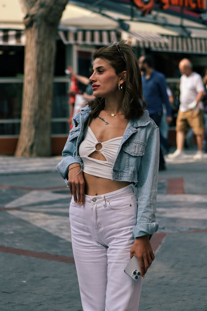Stylish woman wears a jean jacket and holds a phone while standing outdoors on a city sidewalk.